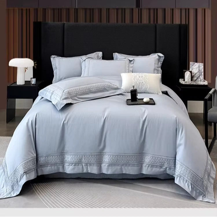 A neatly made bed in a modern bedroom setting, dressed in a light blue-gray bedding set with matching pillows and decorative embroidered borders. The bed is flanked by black bedside tables with lamps and decor, set against a dark wood-paneled wall.