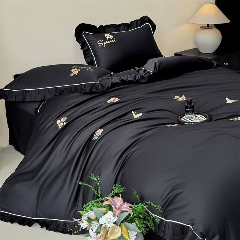 Black satin bedding set with white piping and delicate floral embroidery, styled on a bed in a bedroom setting with a bedside lamp and decorative flowers.