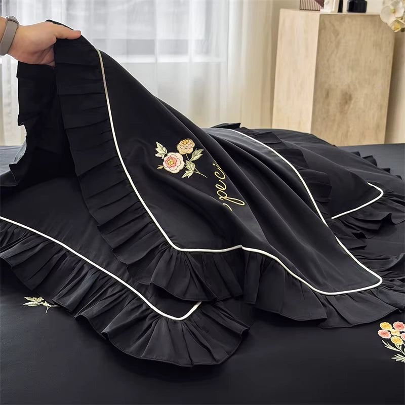 Black bedspread with layered ruffles, white piping, and floral embroidery, being held up by a hand in a bedroom setting.