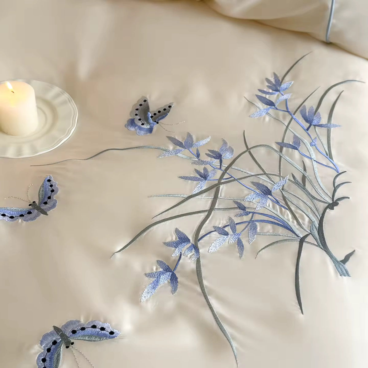 Close-up of a cream satin bedding surface with embroidered blue butterflies and delicate blue-gray floral vines. A lit white candle in a white plate sits at the upper left.