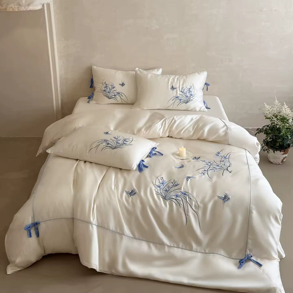 An off-white bedding set with blue floral embroidery and tassel ties, styled on a bed in a softly lit, neutral-toned bedroom.