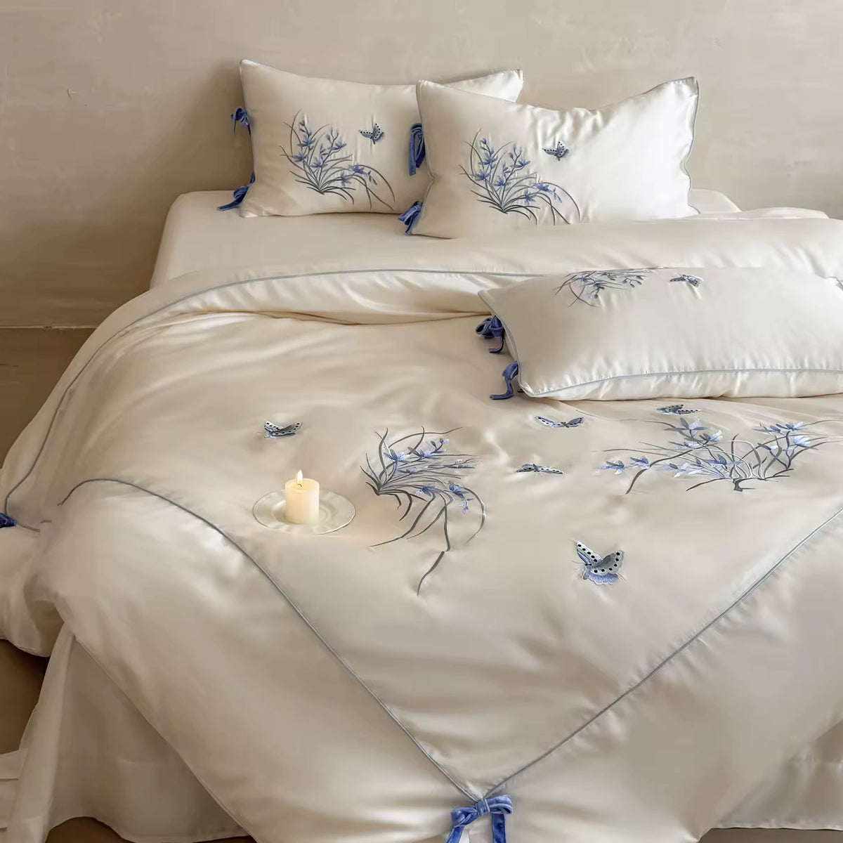 An off-white satin bedding set with blue floral embroidery and butterfly motifs, styled on a bed with matching pillows and a lit candle.
