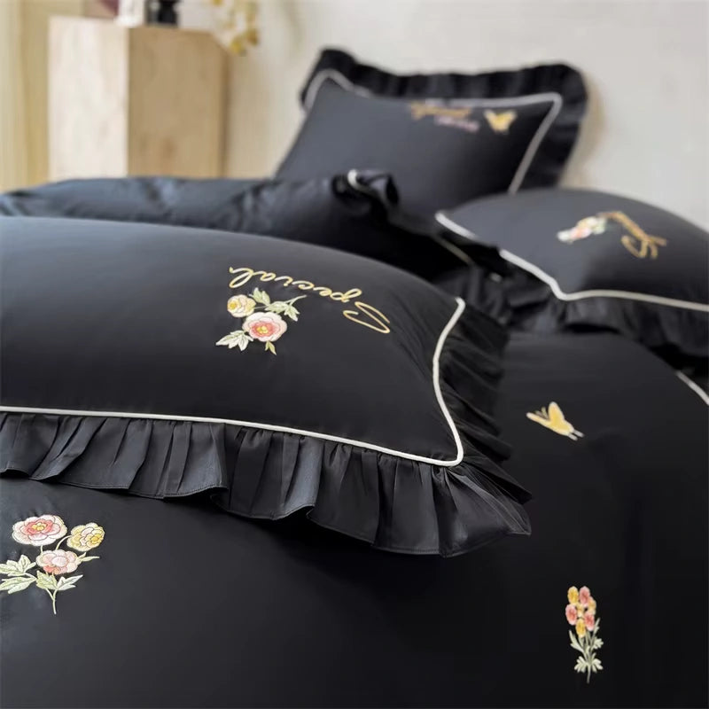 Black bedding set with ruffled pillowcases and duvet cover, featuring gold embroidered script and floral motifs, shown on a bed in a softly lit bedroom.